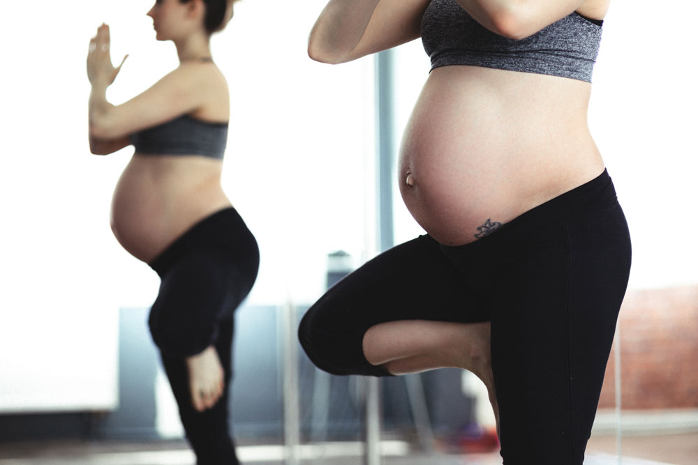 2 pregnant women with bare bumps standing on one leg doing yoga